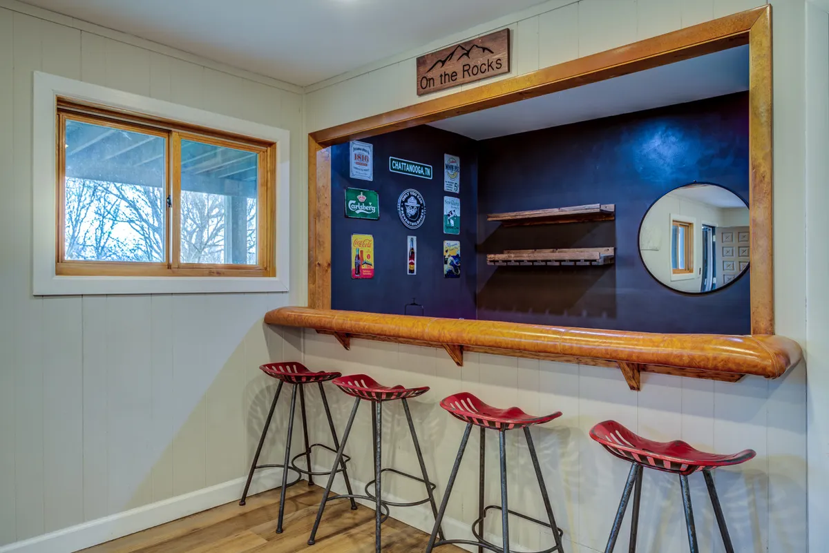 A stylish and inviting bar area features a polished counter with bar stools, illuminated by warm lighting and surrounded by decorative signage. Large windows let in natural light and offer glimpses of the outdoors, while a group of friends enjoy drinks and conversation, evoking a lively, social atmosphere perfect for evening gatherings.