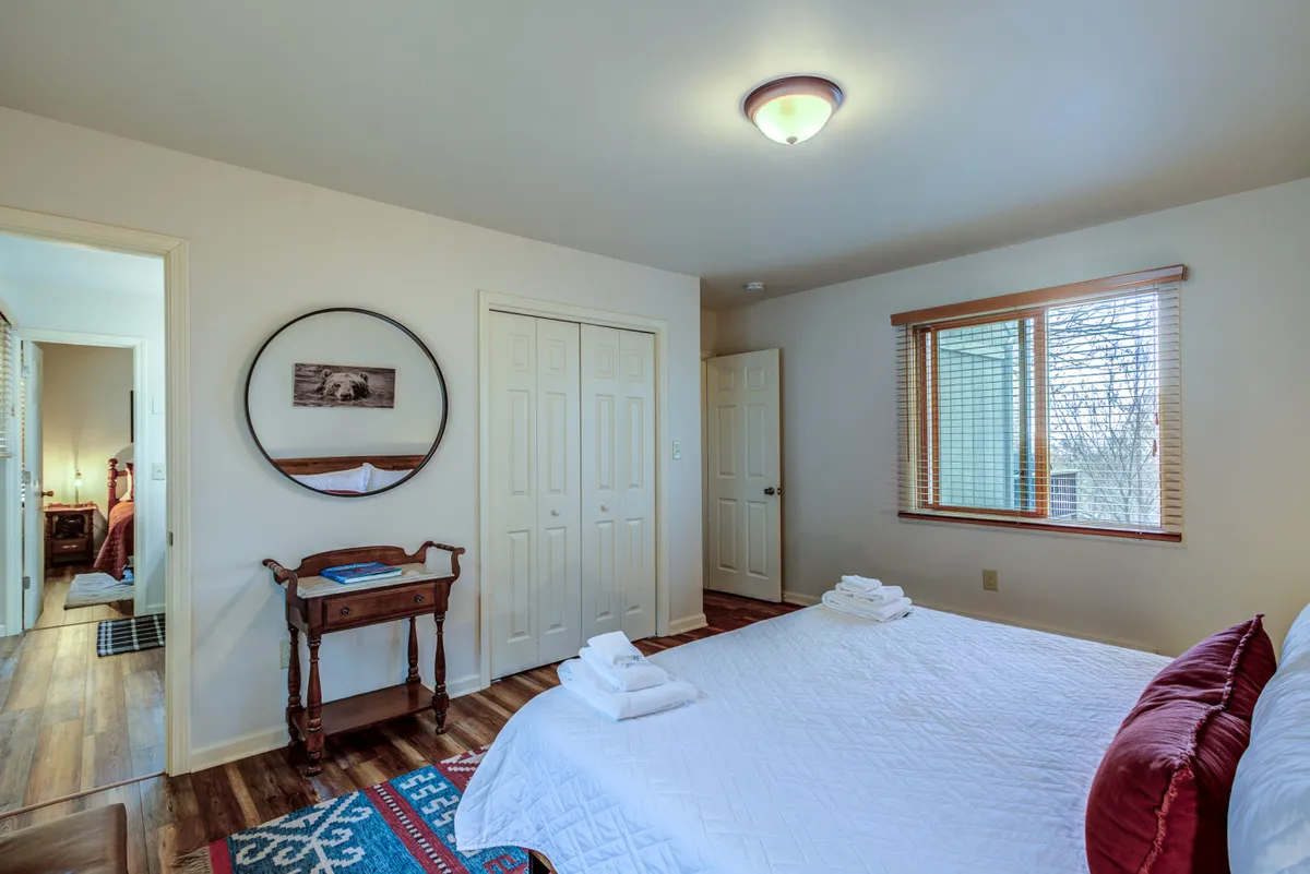 A cozy bedroom with a neatly made bed featuring white linens and decorative pillows, a round mirror on the wall, and a wooden side table. Natural light fills the space, inviting relaxation and rest after an active day.