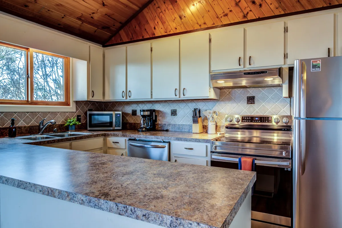 The image showcases a welcoming kitchen with a prominent island, stainless steel appliances, a wooden ceiling, and a large window that likely offers views to the outdoors. The space is filled with natural light, creating an inviting atmosphere for gathering, cooking, and enjoying meals together, especially on days when outdoor exploration is limited by weather.