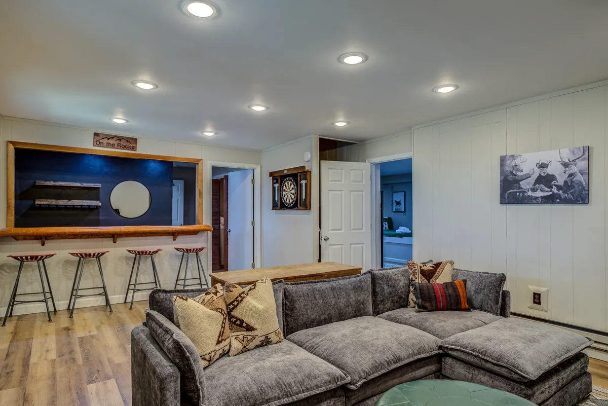 The game room blends comfortable sofa seating with a prominent bar area, dartboard, and vibrant wall art. The setup encourages activities like relaxing, playing games, and socializing, all in a welcoming, casual space ideal for group entertainment.