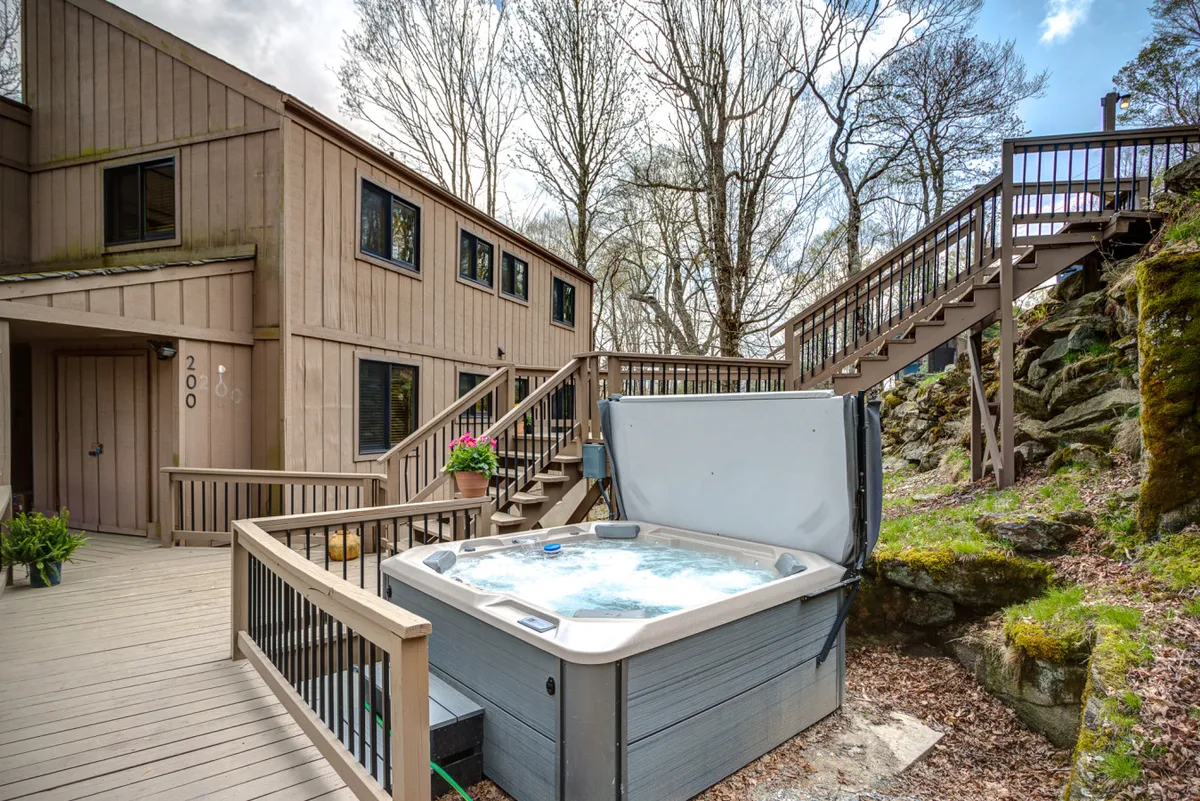 The image showcases the spacious wooden deck of Summit Sky Lodge at sunset, featuring a bubbling hot tub with sweeping mountain views. There are planters bursting with flowers, a staircase leading up to the deck, and a tranquil atmosphere perfect for relaxing or entertaining friends and family.