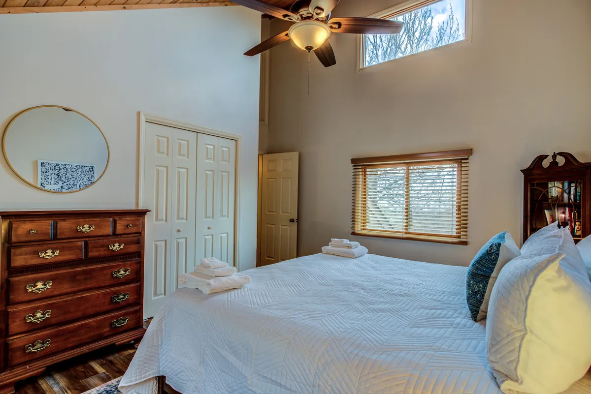 This serene bedroom features a plush bed with crisp white bedding, a wooden dresser, a large mirror, and a window with blinds that allows natural light to flood the space. The view through the window hints at the scenic surroundings, making it easy to imagine morning coffee enjoyed in comfort while overlooking the landscape.