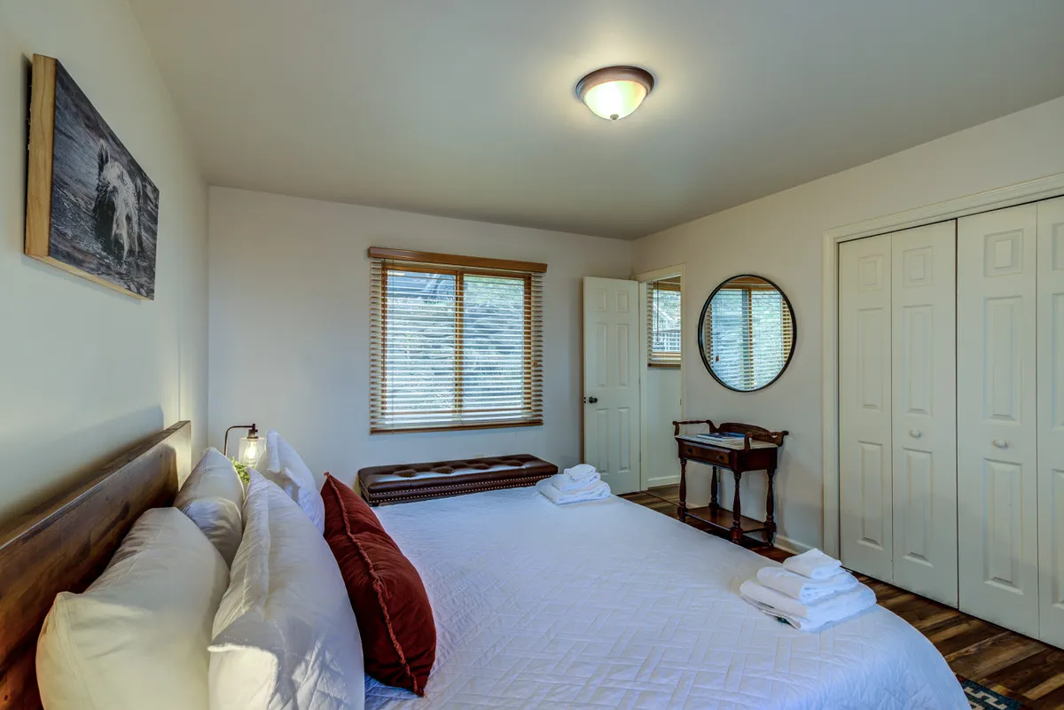 This image features a bed with plush pillows, a wooden bench at the foot of the bed, a round mirror on the wall, and sunlight filtering through a window with blinds. The setup evokes a mood of relaxation and comfort, making it ideal for restful nights.