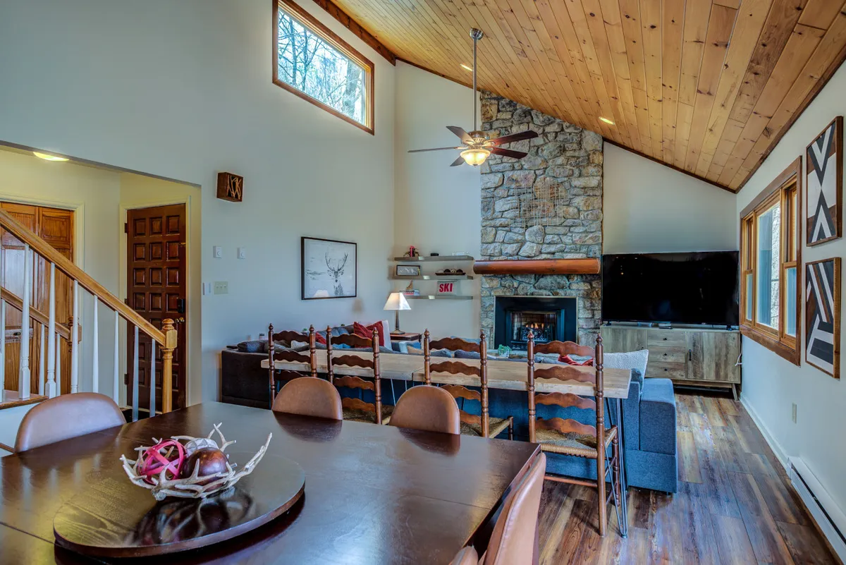 A warm, welcoming interior showcasing a stone fireplace, a large dining table, wooden staircase, and floor-to-ceiling windows framing the surrounding landscape. The space is filled with natural light and arranged to facilitate group interaction and relaxation.