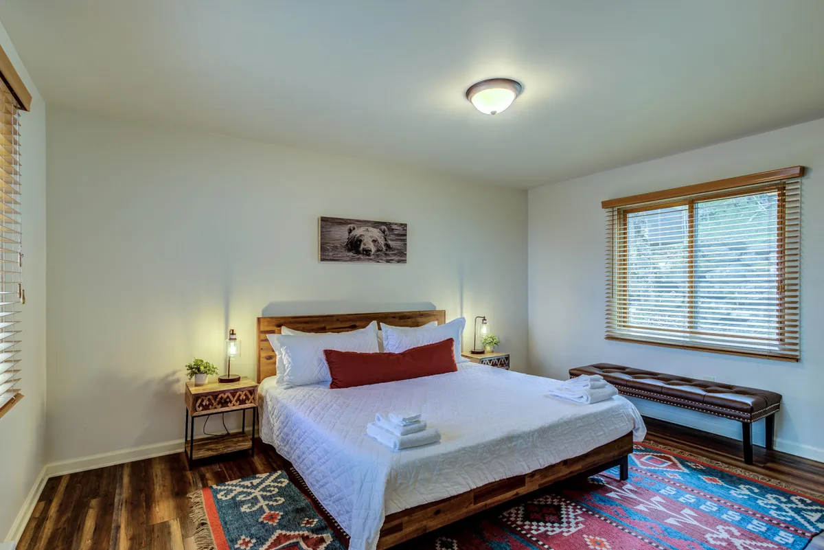 A cozy bedroom with white linens accented by a red pillow, a geometric patterned rug, a wooden bench, and whimsical bear wall art. The window brings in natural daylight and offers a glimpse outside, reinforcing the sense of privacy and comfort.