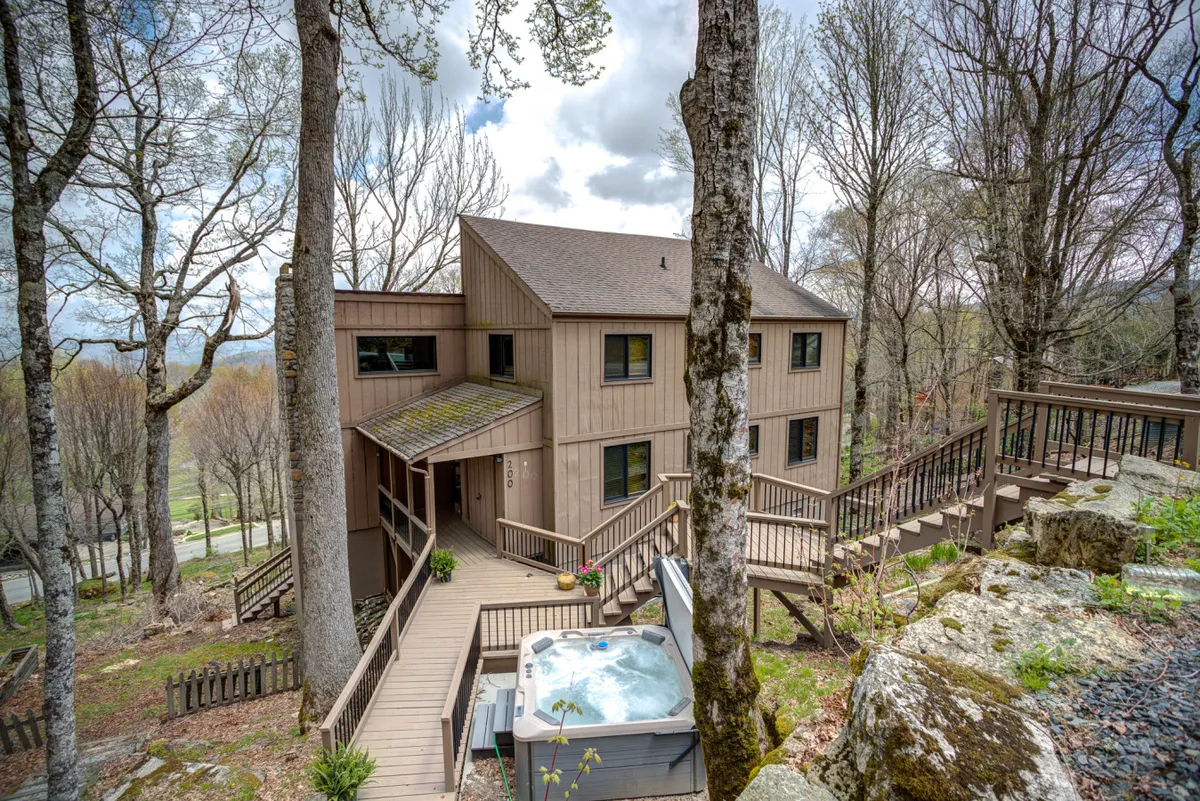 A spacious, wooden deck extends from the house, featuring a bubbling hot tub with views of surrounding trees and rolling mountains. The deck is furnished for comfort and relaxation, inviting guests to gather outdoors regardless of the season.