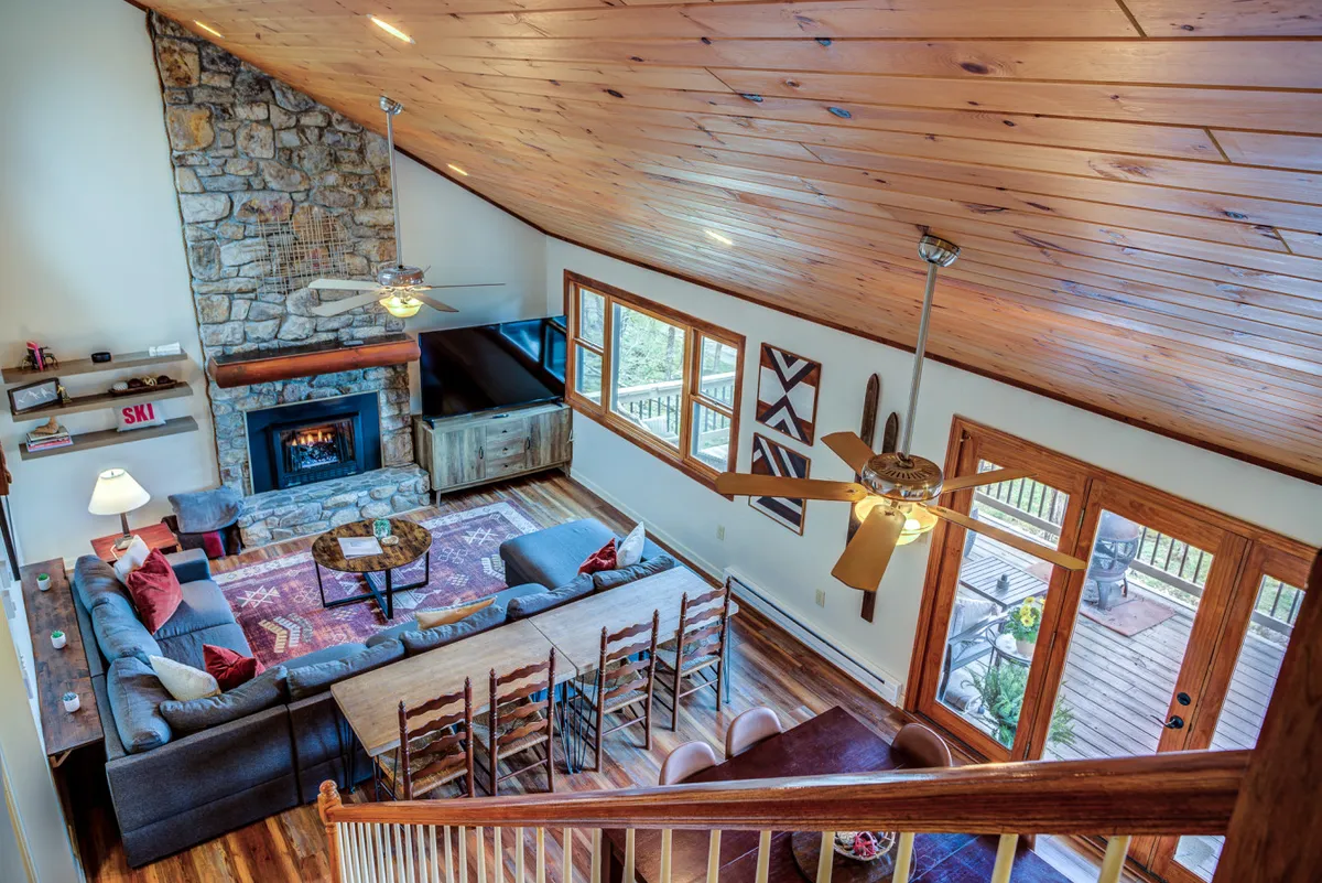 A spacious open living room featuring a stunning stone fireplace, a large wooden dining table that seats a crowd, expansive windows revealing mountain views, and plush, inviting seating. The room is flooded with natural light and exudes warmth and comfort, perfect for group gatherings.