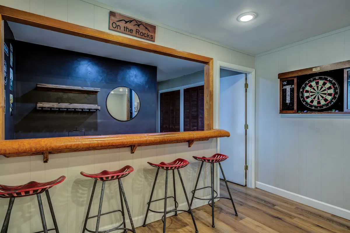 A cozy game room featuring a stylish bar counter, dartboard, round mirror, and the playful 'On the Rocks' sign, all set within a welcoming, social atmosphere. The space suggests opportunities for après-ski fun, lively tournaments, and gatherings.